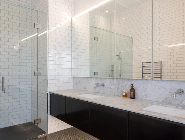 Bright contemporary bathroom with white subway-tiled walls, a glass-enclosed walk-in shower, expansive mirrored wall cabinetry, and a long double vanity. The vanity features dark flat-panel cabinetry, a pale marble benchtop and matching marble splashback, with chrome tapware and a heated towel rail. Small-format dark floor tiles provide contrast.