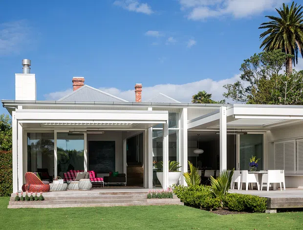 A bright contemporary single-storey home viewed from the landscaped front lawn, featuring a white rendered and metal-clad facade, low-pitched gabled roof, broad glazed living areas, and an expansive covered outdoor entertaining deck. Tropical palms and structured planting surround the house.