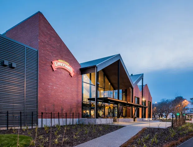 Exterior architectural photograph of a contemporary multi-storey commercial building at dusk. The prominent facade combines red brick volumes, dark horizontal metal cladding, extensive glazing, and angular pitched roof forms. A glazed entrance pavilion projects beneath a deep dark canopy, with landscaped planting beds, pedestrian paths, street lighting, and urban context visible around the building.