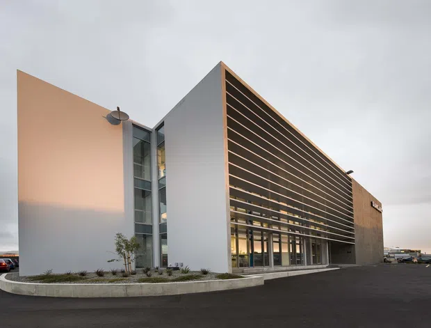 Exterior view of a contemporary commercial building with crisp white and grey wall planes, a tall glazed vertical circulation zone, and a prominent wood-toned horizontal louvred screen over the main glazed facade. The building sits on a paved site with minimal landscaping under an overcast sky.