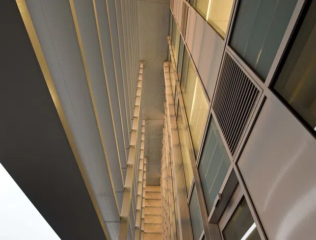 Upward-looking view within a tall contemporary building courtyard or covered external passage. The composition shows repetitive vertical fins and columns, exposed concrete surfaces, extensive glazed panels, metal-framed windows, and projecting upper-level structure. Warm interior lighting reflects in the glazing, while trees and overcast sky are visible at the lower edge.