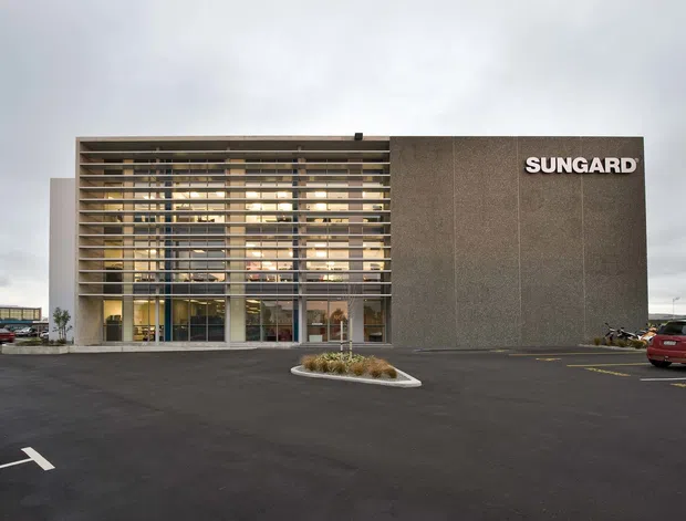 A modern commercial building viewed from a large asphalt parking area. The facade combines a tall glazed entrance volume screened by closely spaced horizontal fins with a solid dark textured wall carrying the SUNGARD sign. Overcast skies and surrounding parked vehicles frame the low-rise commercial setting.