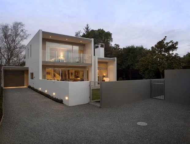 A contemporary two-storey residential house viewed from the front at dusk. The building has crisp white rendered walls, a strong box-like form, large glazed openings with slim dark frames, recessed exterior lighting, a projecting upper-level balcony or glazed terrace, and an attached single-car garage. A broad gravel driveway, front gate, low boundary walls, and mature trees surround the property.