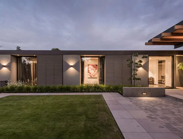 A contemporary single-storey residential facade viewed from a landscaped front courtyard at dusk. The building has a long, low horizontal composition with dark grey rendered wall panels, extensive horizontal timber-look screening, recessed glazed openings, warm wall lighting, and a projecting layered timber soffit or roof overhang at the right. A paved path and driveway-like forecourt lead to the entry, with lawn, planting, a raised concrete planter, and glimpses of a warmly lit interior.
