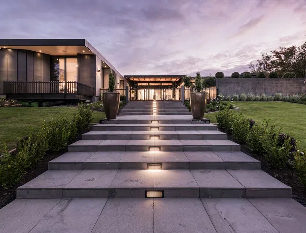 Twilight exterior view of a contemporary single-storey residence approached by a broad flight of pale stone-paved steps with integrated linear lighting. The home has a dark, flat-roofed facade with extensive glazing, a raised timber deck, and a covered pergola or entry pavilion aligned along the central axis. Formal lawns, clipped planting, large planters, and a rendered boundary wall complete the landscaped approach.