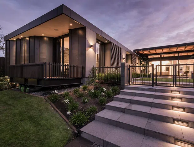 Contemporary single-storey residence photographed at dusk, featuring dark vertical cladding, a projecting flat-roofed living wing, extensive glazing, and a covered outdoor entertaining area connected by broad illuminated tiled steps. Landscaped garden beds and a sheltered courtyard-like approach complete the exterior composition.