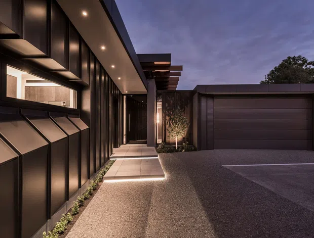 Twilight exterior view of a sleek contemporary residential facade and integrated garage. The dark metal-clad building features projecting angular window surrounds, extensive vertical glazing, recessed soffit lighting, a covered entry walkway, timber-like projecting roof or pergola elements, landscaped planting, and a broad textured driveway.