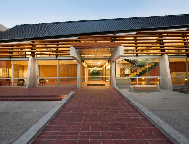 Front exterior view of a substantial modern building with a long dark corrugated metal gable roof, exposed horizontal timber sun-shading slats, concrete structural piers, extensive glazing, warm timber wall panels, and a broad red tiled entry walkway. The central glazed entrance reveals a brightly lit interior with a staircase and planted feature visible beyond.