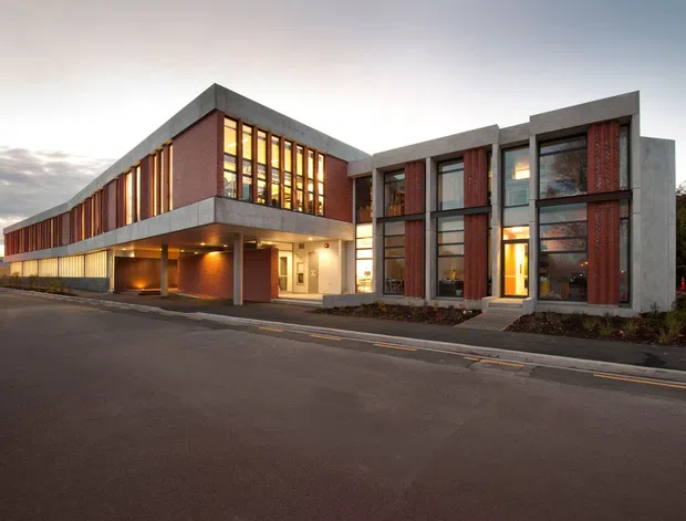 Exterior view of a large contemporary institutional or commercial building at dusk. The complex combines long, low rectangular volumes with projecting upper floors, exposed red brick wall sections, dark metal-framed glazing, and prominent grey concrete frames and slabs. Warm interior lighting glows through the extensive windows, with a paved street and entry forecourt in the foreground.