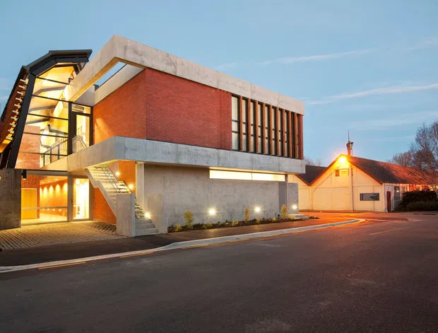 Exterior street view of a contemporary multi-storey building at dusk. The structure features exposed concrete slabs and walls, a prominent red brick upper volume, extensive glazed entry and stairwell areas, vertical timber-like screening, and a sharply angled roof element. A smaller traditional neighboring building is visible to the right, with landscaped planting and paved street frontage.