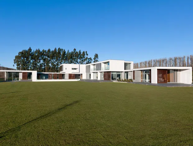 Wide exterior view of a large, low-profile contemporary residence set behind an expansive lawn. The building has crisp white rectangular volumes, extensive floor-to-ceiling glazing, dark timber-clad sections, and multiple connected wings around a sheltered entry or outdoor area. Mature trees form a backdrop under a clear blue sky.