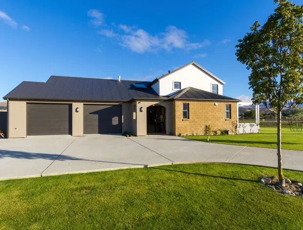 A detached single-storey suburban/rural home viewed from the front, with a dark pitched metal roof, pale rendered upper walls, a warm tan brick entry wing, and an attached double garage. A broad concrete driveway and neatly maintained lawn frame the facade, with distant open landscape visible beyond.