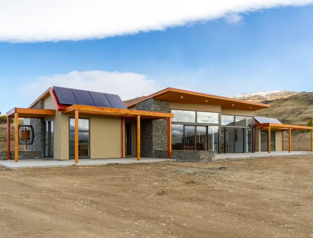 A low-slung contemporary single-storey building set in a rugged, sparsely vegetated landscape. The facade combines large glazed sections, dark stacked-stone walls, warm timber posts and soffits, pale rendered walls, and a pitched roof with dark solar panels. Broad roof overhangs and covered outdoor areas extend around the building.