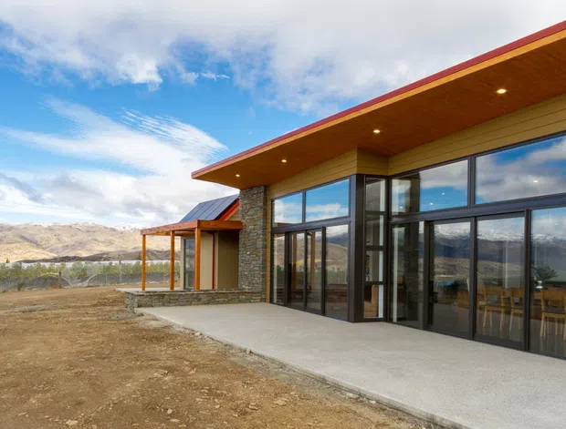 Contemporary single-storey home exterior with expansive dark-framed glazing, a broad timber-lined roof overhang, and a stone-clad feature wall. The building sits on a dry, open site with mountain views and a partially covered outdoor terrace. The facade combines warm timber soffits, natural stone, pale wall cladding, and concrete paving.