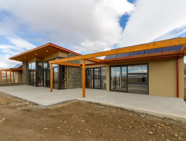 A low-profile contemporary single-storey building set in a rugged, open landscape. The exterior combines warm timber pergola beams and posts, broad floor-to-ceiling glazing with dark frames, smooth beige rendered walls, and low-pitched roof forms. A large concrete terrace runs along the glazed facade, with exposed earth in the foreground and mountainous terrain visible beyond.