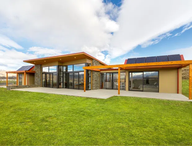 Single-storey contemporary house set in an open grassy landscape, featuring a low-pitched roof with broad timber eaves, extensive floor-to-ceiling glazing, dark stone-clad walls, warm timber posts and beams, and a long covered outdoor terrace. Solar panels are visible on the roof, with rolling hills in the background.