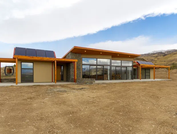 A low-profile contemporary single-storey building set in a rugged, sparsely vegetated mountain landscape. The facade combines dark stone masonry, warm timber-framed verandah and canopy structures, broad floor-to-ceiling glazing, and a projecting flat or gently sloped roof with visible solar panels. The surrounding ground is undeveloped and gravelly, with hills rising behind the building.