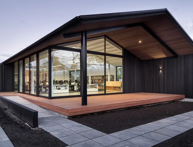 A contemporary single-storey pavilion-like building with a dramatic pitched gable roof, dark vertical cladding, and extensive floor-to-ceiling glazing. A broad timber deck wraps the glazed living or work space, bordered by pale square-paver walkways and dark landscaped beds. Warm timber lining is visible beneath the deep roof overhang, while the interior appears bright and open with shelving, desks, and furniture visible through the glass.
