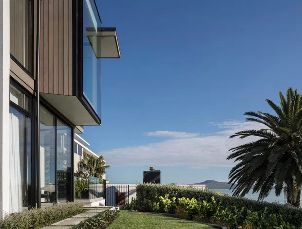 Exterior architectural photograph of a contemporary multi-level residence beside a waterfront or coastal outlook. The building features expansive floor-to-ceiling glazing, dark vertical timber cladding, white rendered walls, projecting balconies or overhangs, and a landscaped lawn with hedges and a mature palm. A railing and distant water with hills are visible beyond the property.