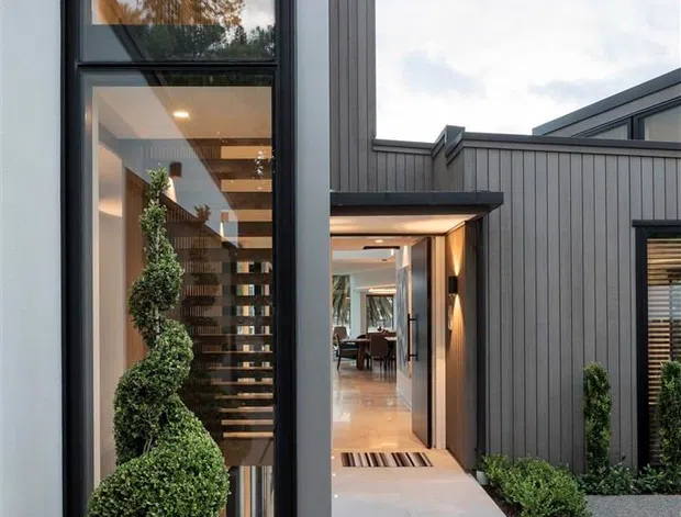 A contemporary residential entry courtyard viewed from outside, featuring a tall glazed entrance volume, dark vertical-clad walls, pale large-format paving, and a recessed front door. Through the glazing and open doorway, a sculptural open-riser staircase, warm timber detailing, and a dining area are visible. Formal clipped topiary and low planting frame the path, while dark textured aggregate paving surrounds the entry.