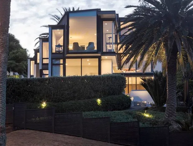Exterior view of a contemporary multi-storey residence at dusk, featuring extensive floor-to-ceiling glazing, dark framed projecting volumes, warm interior lighting, manicured hedges, tall palms, and a dark stone retaining wall along the street frontage.