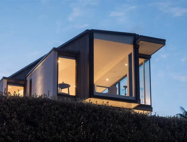 Exterior view of a contemporary multi-level dwelling at dusk, featuring strong geometric volumes, extensive floor-to-ceiling glazing, dark vertical cladding, projecting roof planes, and warm interior lighting. Dense hedging obscures the lower level, while palm vegetation is visible at the edge of the scene.