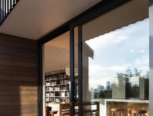 Exterior view toward a contemporary home through large black-framed sliding glass doors. A timber deck and dark horizontal timber cladding frame the opening, with a balcony and black vertical railing overhead. Through the glazing, a warm timber-floored interior is visible with a built-in bookshelf, dining table, and kitchen island or counter with timber stools. The image emphasizes indoor-outdoor connection, expansive glazing, and layered timber finishes.