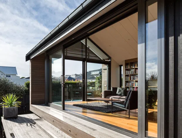 Exterior view of a contemporary residential extension with a low-pitched roof, dark vertical cladding, and extensive floor-to-ceiling glazing. The glazing reveals a warm timber-floored living room with a lounge chair, rug, and built-in bookshelf. A timber deck and planted container sit in the foreground, while neighbouring homes are visible beyond.