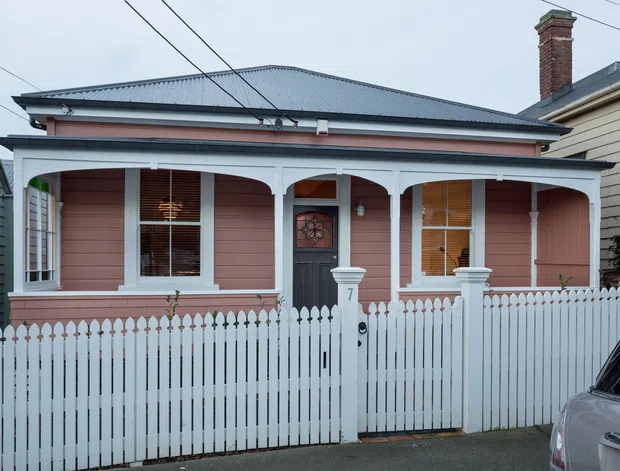 Street-facing view of a single-storey heritage-style timber cottage with a pink weatherboard facade, grey metal hipped roof, decorative white verandah posts and fretwork, enclosed front verandah, brick chimney, and a white picket fence with gate. A neighboring building and part of a parked car are visible at the edges.