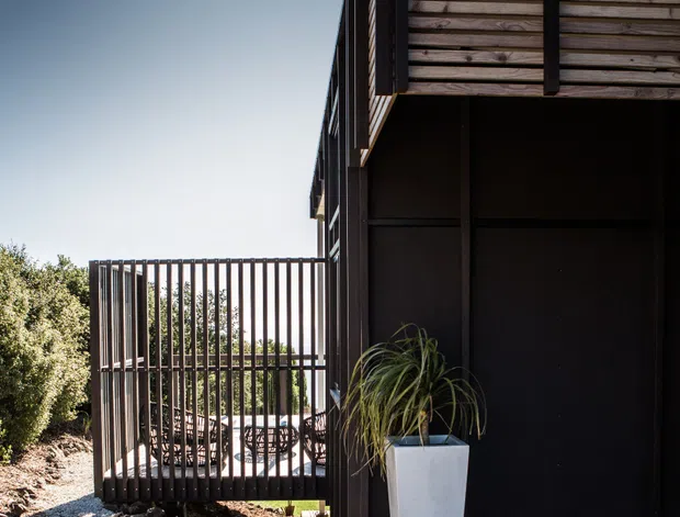 Exterior view of a contemporary dark-clad building with a projecting upper level, horizontal timber screening, and a pale timber deck. A slatted metal privacy fence encloses a small outdoor area, while gravel and dark rock landscaping define the side walkway. The image emphasizes strong geometric lines, shadow, and contrast against a clear sky.