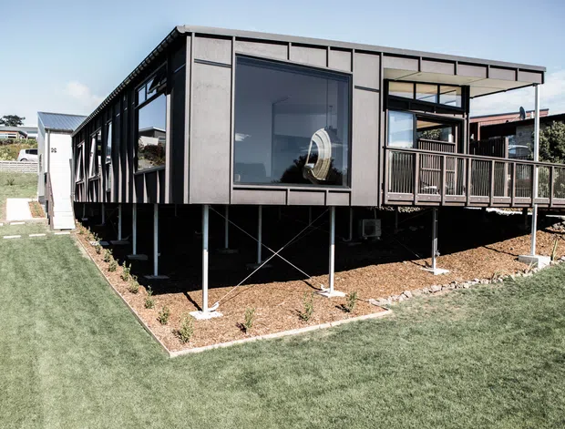 Exterior view of a contemporary elevated modular or prefabricated home with a dark, box-like facade, extensive glazing, and a covered timber deck. The building is raised on slender steel supports with diagonal bracing, surrounded by lawn and gravel landscaping in a suburban setting.