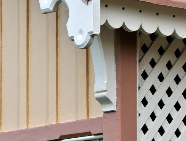 Close-up architectural detail of a painted exterior facade featuring a pink corrugated metal roof, cream vertical weatherboard-style wall panels, white decorative scalloped eaves, ornate brackets, and white diagonal lattice screens. The image emphasizes decorative period detailing and layered trim rather than a complete room or building.