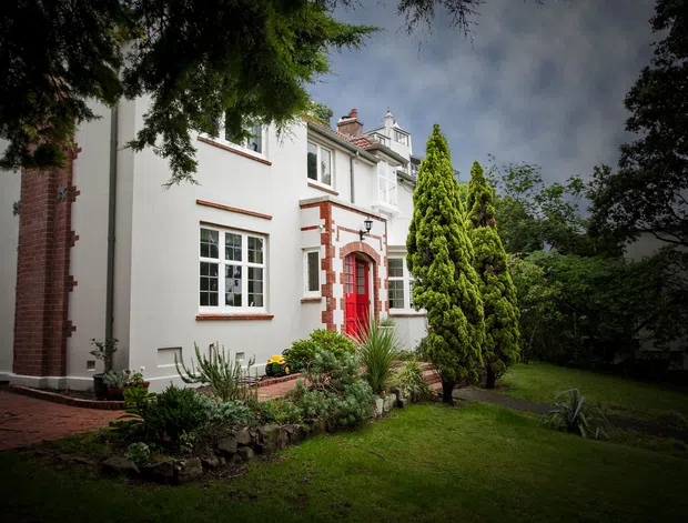 Exterior view of a substantial period-style residential home set within a landscaped garden. The rendered facade has contrasting red brick detailing, a red feature entrance with arched surround, white-framed multi-pane windows, decorative upper-level trim, and chimney stacks. Mature trees, lawn, planted beds, and a brick path frame the building.