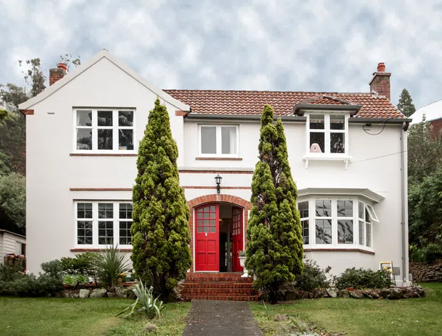 Front exterior view of a two-storey white rendered house with a symmetrical gabled central section, tiled low-pitched roof, red front door, multi-pane timber windows, brick chimneys, and a projecting bay window. Brick steps and a planted front garden lead to the entry.