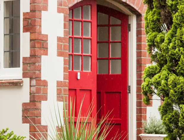 Close-up exterior view of a traditional white-rendered building facade with red brick quoins, decorative brick banding, and a brick-arched entrance. The entrance has a vivid red glazed timber door beneath a black wall-mounted lantern. Brick steps and landscaped planting, including clipped evergreen shrubs and spiky ornamental grasses, frame the entry.