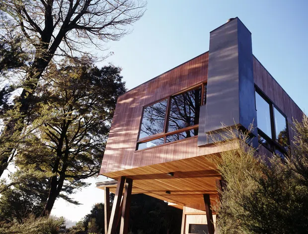 Exterior view of a contemporary elevated building surrounded by mature trees and landscaping. The structure features a sharply angular, box-like form, extensive dark-framed glazing, warm vertical timber cladding, a timber-lined soffit, and a prominent tall chimney or service tower. The ground level is open beneath the upper volume, supported by angled posts.