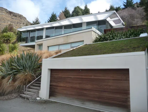 Exterior view of a contemporary multi-level hillside residence with a large integrated garage in the foreground. The house has a long low-pitched roof with a prominent glazed upper level, light rendered or masonry walls, horizontal glazing, and a timber garage door. A planted roof or landscaped terrace slopes above the garage, with native-style planting and exposed rocky terrain surrounding the property.