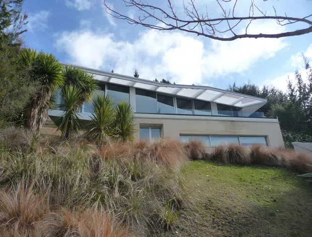 A modern two-storey residence set into a landscaped, sloping site. The home has a long horizontal composition, extensive upper-level glazing, a projecting roof with visible metal roofing, and pale rendered walls. Native-style grasses and spiky plants surround the building, with mature vegetation behind it.