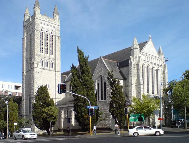 Exterior photograph of a substantial historic Gothic Revival church on an urban street corner. The building has pale grey masonry walls, a tall square bell tower with corner pinnacles, steep gabled roofs, pointed-arch windows, decorative tracery, and a large nave or transept-like volume. Mature trees and traffic signals line the foreground, with surrounding urban buildings visible behind.