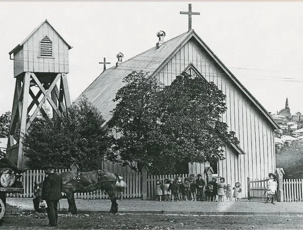 Black-and-white historical photograph of a small timber church with steep gabled roofs, vertical weatherboard cladding, decorative gable detailing, and multiple crosses. A tall timber bell tower stands beside the church, with a horse-drawn cart and people outside behind a picket fence. Urban buildings are visible in the background.