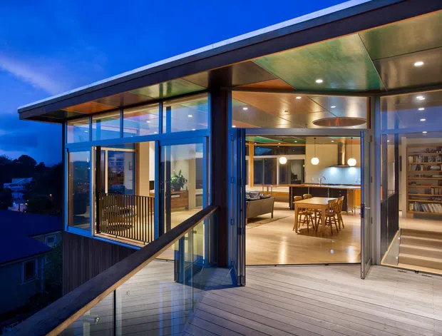 Twilight exterior view of a contemporary elevated home with extensive floor-to-ceiling glazing, a broad low-pitched roof, timber-lined ceilings, dark structural framing, and a generous timber deck. The open interior reveals a dining area, kitchen, living space, and tall built-in shelving, with a stair descending at the right. The home appears positioned on a sloped suburban site.