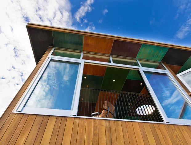 Low-angle exterior view of a contemporary building facade with extensive white-framed glazing, timber-clad walls, and a projecting eave lined with multicoloured timber or metal panels. An upper-level interior is visible through the open glazing, including a ceiling, railing, and furniture. Bright blue sky and clouds surround the structure.