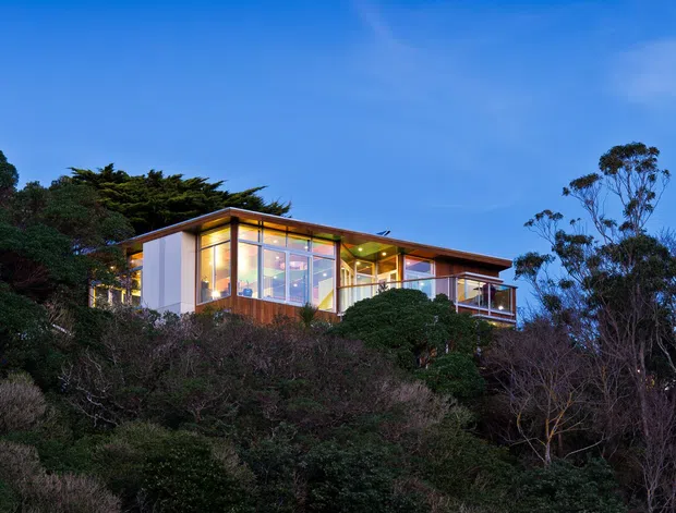 Dusk exterior view of a modern, elevated house set among dense vegetation on a steep, tree-covered site. The dwelling has extensive floor-to-ceiling glazing, timber-toned framing and cladding, a low-pitched roof with broad eaves, and a projecting balcony or terrace. Warm interior lighting contrasts with the deep blue sky and dark landscaped surroundings.