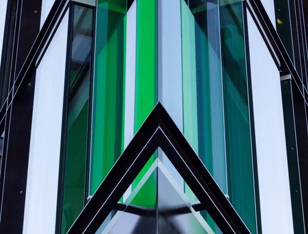 Close-up upward view of a striking modern building facade with sharply angled triangular geometry, black structural framing, extensive glazing, and vivid green, turquoise, blue, and magenta glass panels. The composition emphasizes the dramatic pointed entrance or canopy and vertical facade articulation.