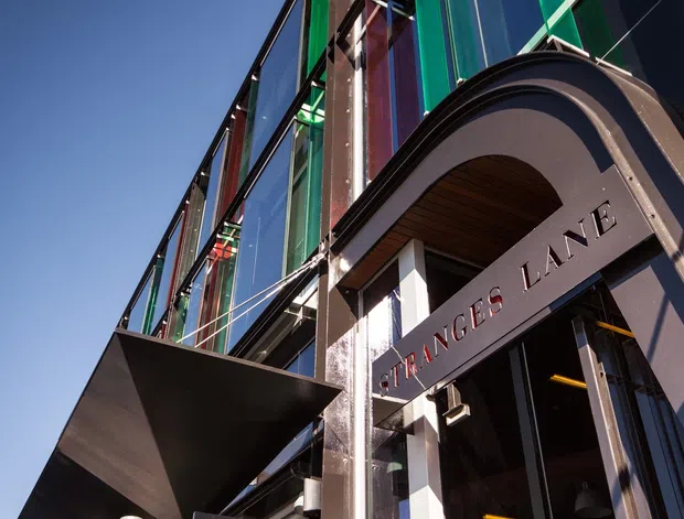 Upward-looking exterior architectural photograph of a contemporary multi-storey commercial building entrance. The facade combines reflective and vividly coloured green, teal, and red glazing with dark metal framing, projecting angular canopies, and a prominent arched entrance surround. The building name “FRANCES LANE” is integrated into the entrance facade, with timber lining visible beneath the arch.
