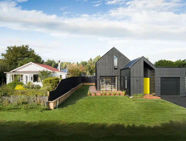 Exterior view of a contemporary dark-clad gabled house with a tall glazed entry, bright yellow front door, and attached single-car garage. The home sits beside a smaller traditional light-coloured house, with front lawn, garden planting, fencing, and a paved driveway visible under a bright sky.