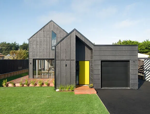 Front exterior view of a contemporary dark-clad residence with two intersecting gabled forms, large glazed openings, a vivid yellow entry door, and an integrated single-car garage. The house is set behind a manicured lawn with timber decking, planting beds, fencing, and a dark asphalt driveway.