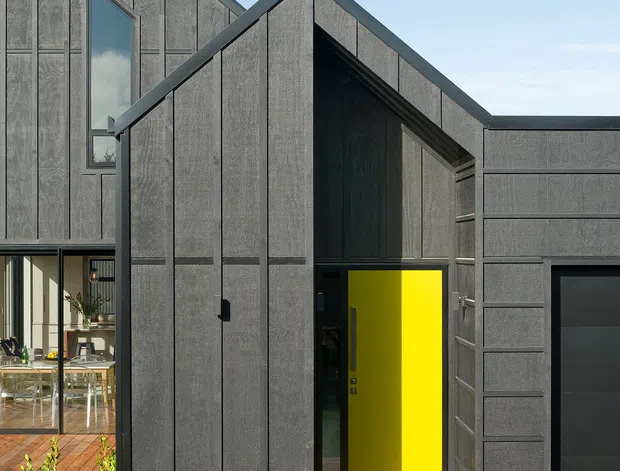Exterior view of a contemporary multi-volume house with steep gabled forms, dark vertically panelled cladding, a vivid yellow front door, and a timber entry deck. A glazed living area and landscaped lawn are visible beside the main entry, with an integrated dark garage volume to the right.