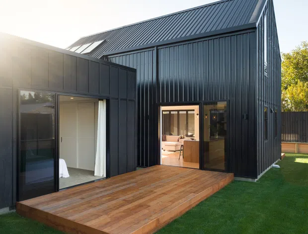 Exterior view of a contemporary dark-clad house or extension with a steep gabled corrugated metal roof, large glazed openings, and a raised timber deck leading to the lawn. The building has a strong black/charcoal palette, crisp geometric form, and an open-plan interior visible through the doors.