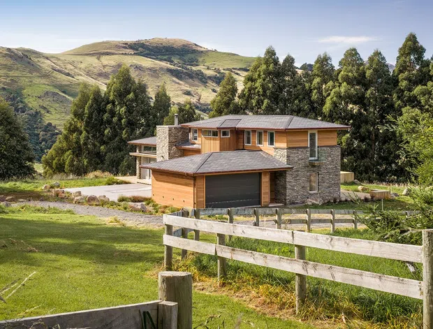 A contemporary two-storey timber-and-stone house set in a rural mountain landscape. The home has a low-pitched roof, warm horizontal timber cladding, substantial stacked-stone walls and chimney, and an attached garage. A timber post-and-rail fence and landscaped lawn occupy the foreground, with wooded hills behind.